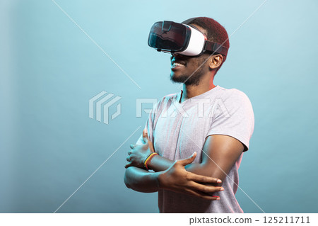 Young male individual enjoying an immersive VR experience, interacting with a 3D simulation. Portrait of african american tech enthusiast wearing virtual reality goggles, stands with crossed arms. 125211711