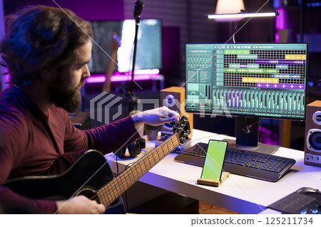 Sound engineer learns tuning acoustic guitar in home studio, watching a video tutorial on internet via mobile phone with greenscreen. Artist learning to play instrument and create tunes. Sound engineer learns tuning acoustic guitar in home studio, watching a video tutorial on internet via mobile phone with greenscreen. Artist learning to play instrument and create tunes. 125211734