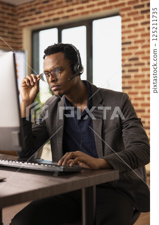 Pensive black man adjusting his glasses and working at desktop pc, focused on client information. Concentrated call center agent wearing headset, attending to customer support tasks on his computer. Pensive black man adjusting his glasses and working at desktop pc, focused on client information. Concentrated call center agent wearing headset, attending to customer support tasks on his computer. 125211735