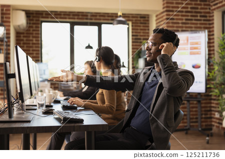 African American telemarketer with headset, pointing at screen with frustration, handling incoming customer requests. Black man working in office, surrounded by technology and communication devices. 125211736