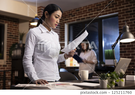 Focused employee reading a document in hand, standing at her office desk during her night shift. Asian corporate manager on her feet, working late, planning a brand strategy for startup company. Focused employee reading a document in hand, standing at her office desk during her night shift. Asian corporate manager on her feet, working late, planning a brand strategy for startup company. 125211760
