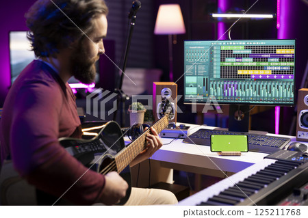Young artist playing acoustic instrument accords in home studio with greenscreen layout shown on smartphone. Singer practices songs and tunes on guitar with strings, watching tutorials. 125211768