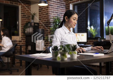 Professional woman working late at night in a modern office, seated at desk, focused on her laptop. Asian female entrepreneur using her personal computer, checking emails and managing business tasks. 125211787