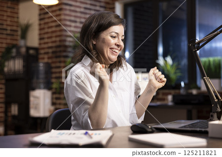 Professional woman celebrates career milestone at desk, dancing in her chair with arms raised, smiling as she looks at her laptop. Excited female entrepreneur feeling triumphant after an achievement. Professional woman celebrates career milestone at desk, dancing in her chair with arms raised, smiling as she looks at her laptop. Excited female entrepreneur feeling triumphant after an achievement. 125211821