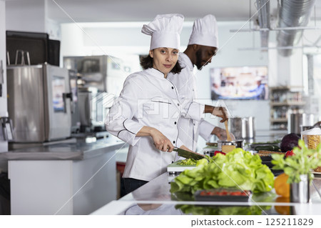 Chef chopping vegetables on a cutting board and preparing ingredients in a modern commercial kitchen. Woman cooking flavorful recipes and fine dining gourmet dishes, chef life. 125211829