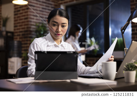 Dedicated female professional working late in corporate office, reviewing business reports on a tablet. Asian manager surrounded with digital devices on her desk, focused on project planning at night. 125211831