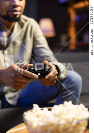Black player enjoying shooter competition with popcorn at home, participating in online video games tournament with friends during game night. Man having fun with championship. Close up. 125211854