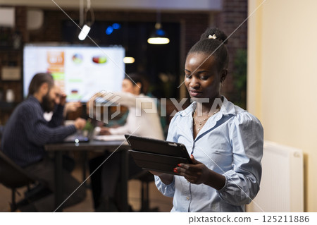 Professional black woman checks financial reports on her tablet while colleagues discuss business strategies. The modern office, late night setting and teamwork highlight productivity Professional black woman checks financial reports on her tablet while colleagues discuss business strategies. The modern office, late night setting and teamwork highlight productivity 125211886