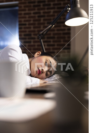 Selective focus of female employee lying asleep at her desk during her night shift. The office is quiet, and her laptop remains open as she rests, trying to recuperate from a demanding workday. Selective focus of female employee lying asleep at her desk during her night shift. The office is quiet, and her laptop remains open as she rests, trying to recuperate from a demanding workday. 125211913