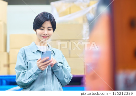 Female businessman in work clothes operating a smartphone in a warehouse Female businessman in work clothes operating a smartphone in a warehouse 125212389
