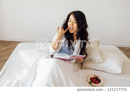 An attractive young Asian woman is lying in bed reading a book and eating berries. A charming female Korean is resting on the sofa in the bedroom. An attractive young Asian woman is lying in bed reading a book and eating berries. A charming female Korean is resting on the sofa in the bedroom. 125217735