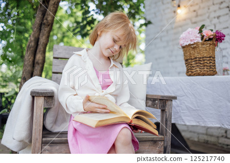 Cute little girl is sitting on wooden chair and looking at book. Pretty child in pink dress and white shirt is leafing through book in garden or in backyard of house. 125217740