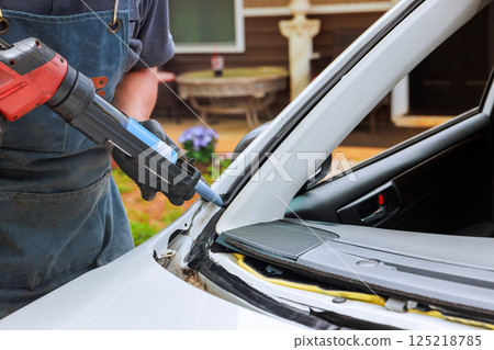 Technician works vehicle trim using caulking gun to seal windshield on car in garage setting, repair process. Technician works vehicle trim using caulking gun to seal windshield on car in garage setting, repair process. 125218785