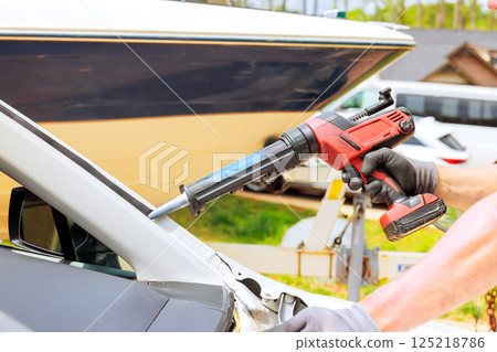 Worker uses caulking gun to repair vehicle windshield beside parked boat Worker uses caulking gun to repair vehicle windshield beside parked boat 125218786