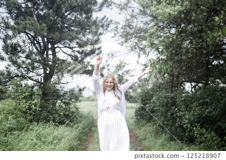Portrait of a laughing young woman in a white dress in the rain. A wet blonde is having fun and dancing in the rain in the park. 125218907