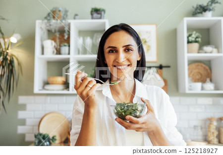Attractive young woman is eating salad of greens in sunny kitchen at home. Smiling brunette girl in white shirt holds fork and transparent bowl with greenery. Attractive young woman is eating salad of greens in sunny kitchen at home. Smiling brunette girl in white shirt holds fork and transparent bowl with greenery. 125218927