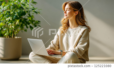Young woman sitting on the floor with a laptop, working or relaxing in a cozy room with natural light streaming in. A large indoor plant adds to the peaceful atmosphere. Concept of remote work and cal 125221080
