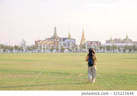 Traveler asian woman in her 30s, backpack slung over her shoulder, explores Wat Pra Kaew with childlike wonder. Sunlight dances on the golden rooftops, and a carefree smile 125221646