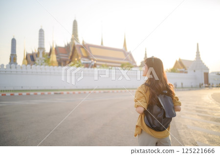 Traveler asian woman in her 30s, backpack slung over her shoulder, explores the intricate details of Wat Pra Kaew with childlike wonder. Sunlight dances on the golden rooftops. Traveler asian woman in her 30s, backpack slung over her shoulder, explores the intricate details of Wat Pra Kaew with childlike wonder. Sunlight dances on the golden rooftops. 125221666