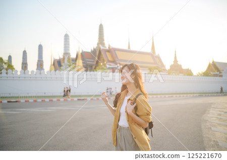 Traveler asian woman in her 30s, backpack slung over her shoulder, explores the intricate details of Wat Pra Kaew with childlike wonder. Sunlight dances on the golden rooftops. 125221670