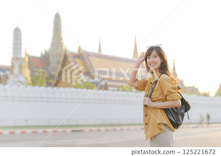 Traveler asian woman in her 30s, backpack slung over her shoulder, explores the intricate details of Wat Pra Kaew with childlike wonder. Sunlight dances on the golden rooftops. 125221672