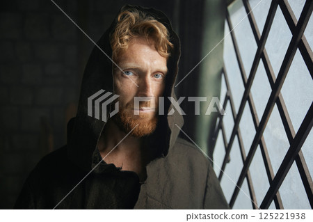 Portrait of pensive monk in medieval costume with hood looking through window of monastery 125221938