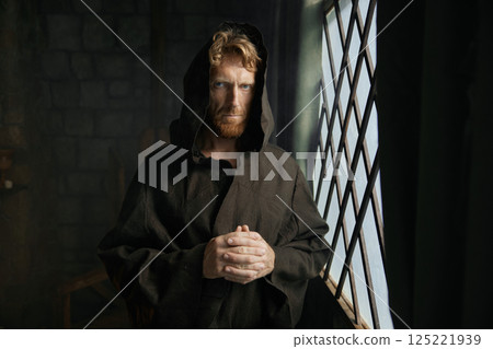 Pensive monk in medieval costume with hood looking through window of monastery 125221939