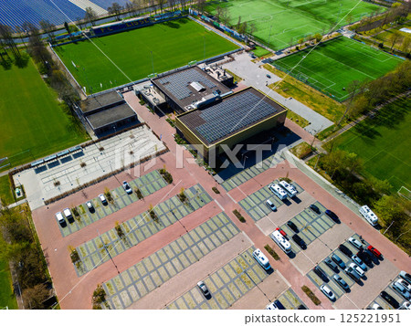 Aerial view of a sports complex with multiple football fields, a track and field stadium, tennis courts, and parking area, surrounded by lush forest and colorful tulip fields in the distance. Aerial view of a sports complex with multiple football fields, a track and field stadium, tennis courts, and parking area, surrounded by lush forest and colorful tulip fields in the distance. 125221951