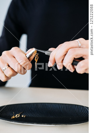Close-up of jewelry maker assembling a gold earring shaped like a dancer using precision pliers. Custom jewelry design, slow fashion, artistic craftsmanship, mindful creation 125222088