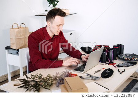 Young adult jewelry maker working at a laptop surrounded by tools and eco-friendly packaging. Handmade crafts economy, sustainable small business, digital retail, creative entrepreneurship 125222097