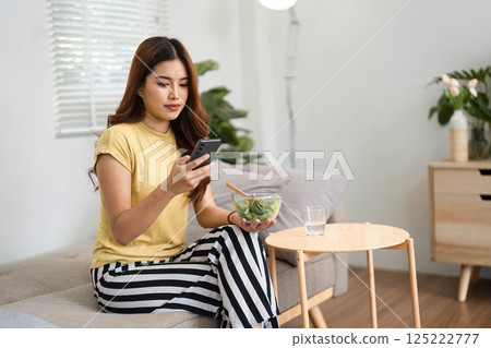 Morning Routine and Healthy Habits. A young woman enjoying a salad while using her phone, embracing healthy living in a cozy setting. 125222777