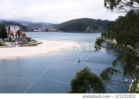 A view of the Ribadesella`s beach, estuary of the river Silla, green hills, mountains, green buoy and a promenade along houses with red tiled roofs 125222852