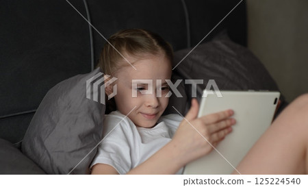 child playing tablet, little girl playing tablet, Girl using tablet while lying in bed relaxing comfortably, Happy child watching cartoon on tablet under gray soft pillows, Young girl smiling at child playing tablet, little girl playing tablet, Girl using tablet while lying in bed relaxing comfortably, Happy child watching cartoon on tablet under gray soft pillows, Young girl smiling at 125224540