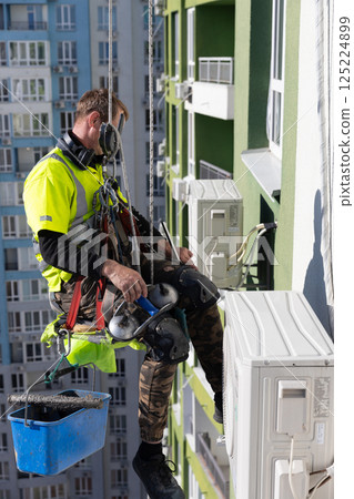 Professional Window Cleaner Performing Task on High-Rise Building with Safety Gear in Bright Yellow Shirt and Harness 125224899