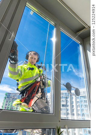 A Professional Window Cleaner in Safety Gear Working at Height to Clean a High-Rise Building Window with a Clear Blue Sky in the Background A Professional Window Cleaner in Safety Gear Working at Height to Clean a High-Rise Building Window with a Clear Blue Sky in the Background 125224914