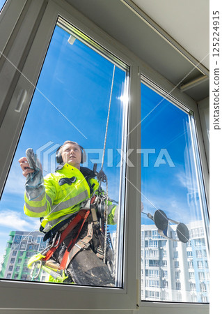 Professional Window Cleaner Performing Exterior Cleaning on High-Rise Building with Clear Blue Sky in Background, Wearing Safety Gear and Harness, Cityscape Visible in Distance Professional Window Cleaner Performing Exterior Cleaning on High-Rise Building with Clear Blue Sky in Background, Wearing Safety Gear and Harness, Cityscape Visible in Distance 125224915