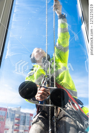 Professional Window Cleaner Man Cleans High-Rise Building Windows with Safety Harness and Equipment in Bright Blue Sky Environment 125225019