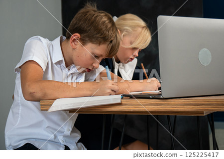 Students Schoolwork Laptop - Two children in school uniform are writing at a table with a laptop. Students Schoolwork Laptop - Two children in school uniform are writing at a table with a laptop. 125225417