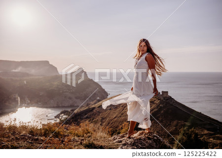 A woman in a white dress stands on a hill overlooking the ocean. The scene is serene and peaceful, with the woman's dress billowing in the wind. The combination of the ocean. 125225422