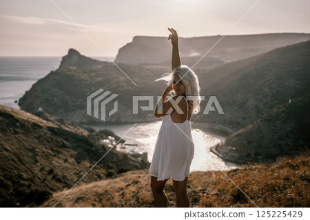 A woman stands on a hill overlooking a body of water. She is wearing a white dress and she is enjoying the view. A woman stands on a hill overlooking a body of water. She is wearing a white dress and she is enjoying the view. 125225429