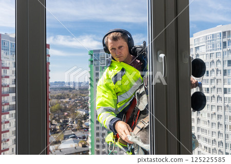 Window Cleaner at High Rise Building: A Dedicated Worker in Safety Gear Performing His Job with Precision from an Elevated Height Overlooking the Cityscape Below Window Cleaner at High Rise Building: A Dedicated Worker in Safety Gear Performing His Job with Precision from an Elevated Height Overlooking the Cityscape Below 125225585
