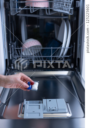 A Close-up View of a Hand Holding Dishwasher Detergent Pack While Preparing to Load the Dishwasher with Dishes Inside, Showcasing the Loading Process and Organization 125225601