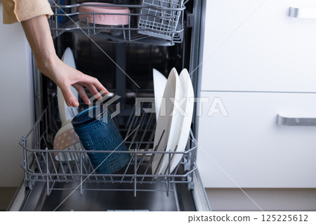 A Hand Reaching for a Blue Glass Jar Inside a Dishwasher Among Clean Dishes: A Moment Captured in Kitchen Cleanliness and Modern Living 125225612