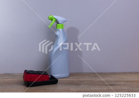 A Cleaning Setup: A Sprayer Bottle and Sponge for Household Cleaning Tasks on a Wooden Surface Against a Minimalistic Gray Wall Background 125225633