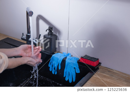 Hands Under Running Water at a Kitchen Sink with Cleaning Supplies Nearby Including Foam Sponge and Rubber Gloves for Cleaning Tasks Hands Under Running Water at a Kitchen Sink with Cleaning Supplies Nearby Including Foam Sponge and Rubber Gloves for Cleaning Tasks 125225636