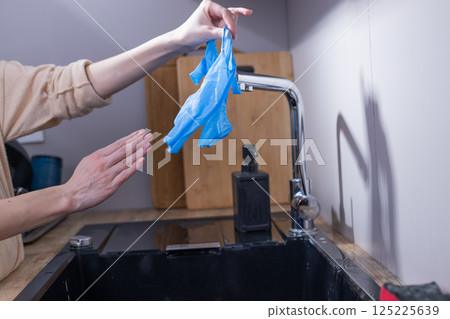 A Person in a Kitchen Washing Disposable Blue Gloves Under Water from a Faucet, Emphasizing Hygiene and Safety in Daily Cleaning Activities 125225639