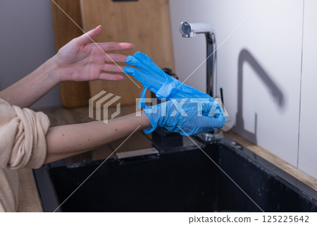 Person Carefully Putting on Blue Disposable Gloves in a Modern Kitchen Sink Setting, Emphasizing Hygiene and Safety Practices during Food Preparation or Cleaning Tasks 125225642