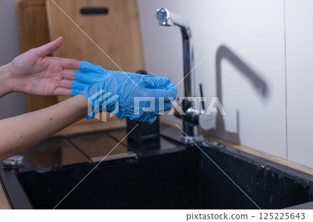 Hands Donning Blue Disposable Gloves While Preparing to Clean in a Modern Kitchen Environment with a Sink and Wooden Elements in the Background 125225643