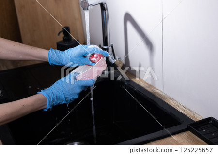 Hands in Blue Gloves Clean a Pet Bowl in a Modern Kitchen Sink with Soapy Water Running from the Faucet, Focus on Hygiene and Care for Pets in Domestic Settings 125225657