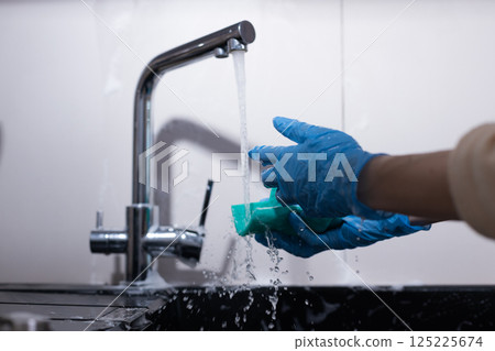 A Person Wearing Blue Gloves Washing Dishes at a Sink, with Soap and Water Flowing from the Faucet in a Brightly Lit Kitchen Environment, Illustrating Cleanliness and Hygiene 125225674